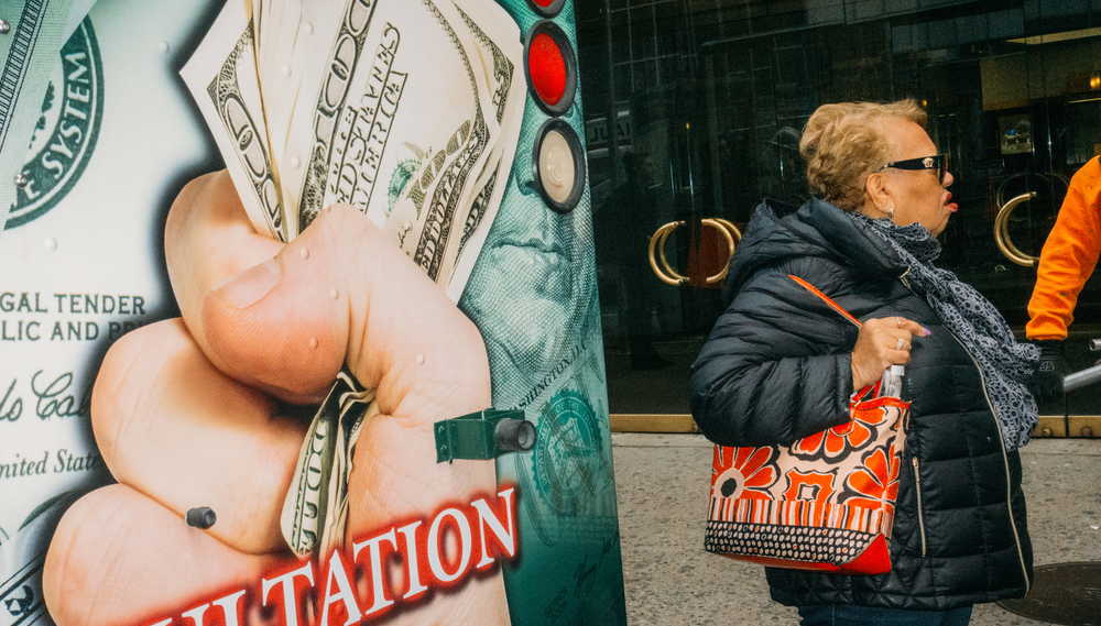 A woman stands to the right of a wall displaying an image of a hand holding several $100 USD bills