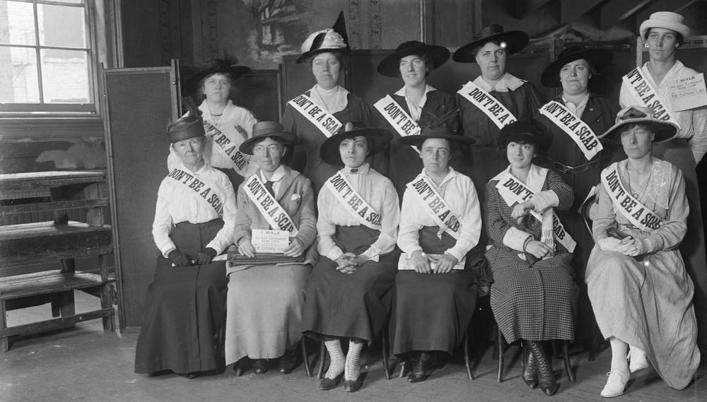 Women delegates aiding striking auto workers seated in two rows wearing sashes that read "DON'T BE A SCAB," circa 1916