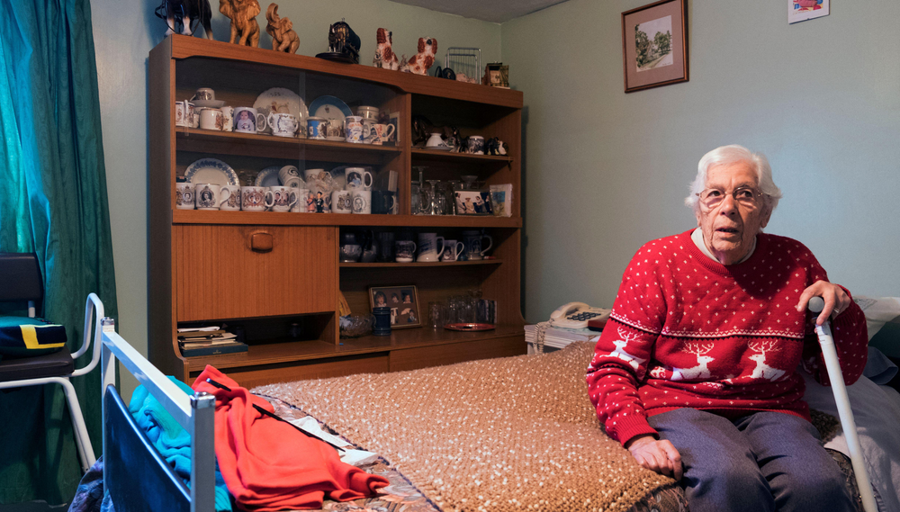 An elderly man sits on the edge of a bed holding a cane