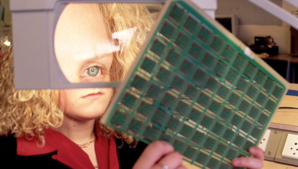 A woman holds and inspects a circuit board in front of a magnifying glass; the magnifying glass magnifies her eye