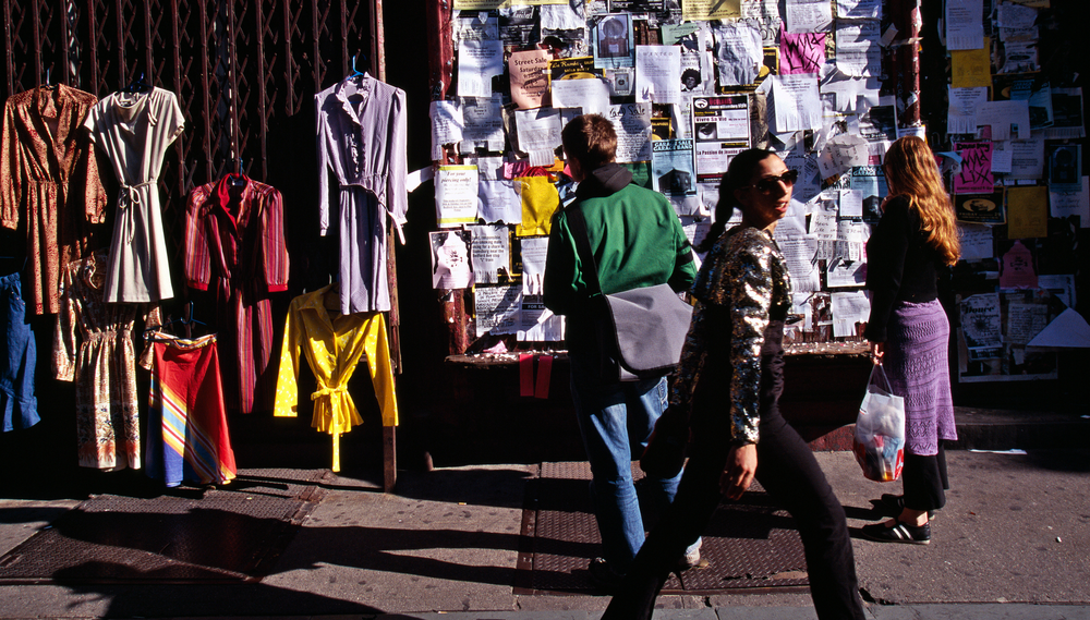 A man and a woman look at a wall of flyers on a Brooklyn sidewalk next to a wall displaying clothes for sale as another woman walks by