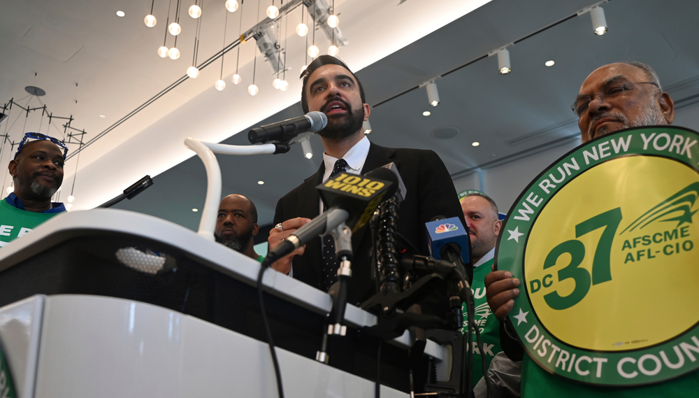 Zohran Mamdani speaks at a podium next to a representative from DC37 holding up a union sign