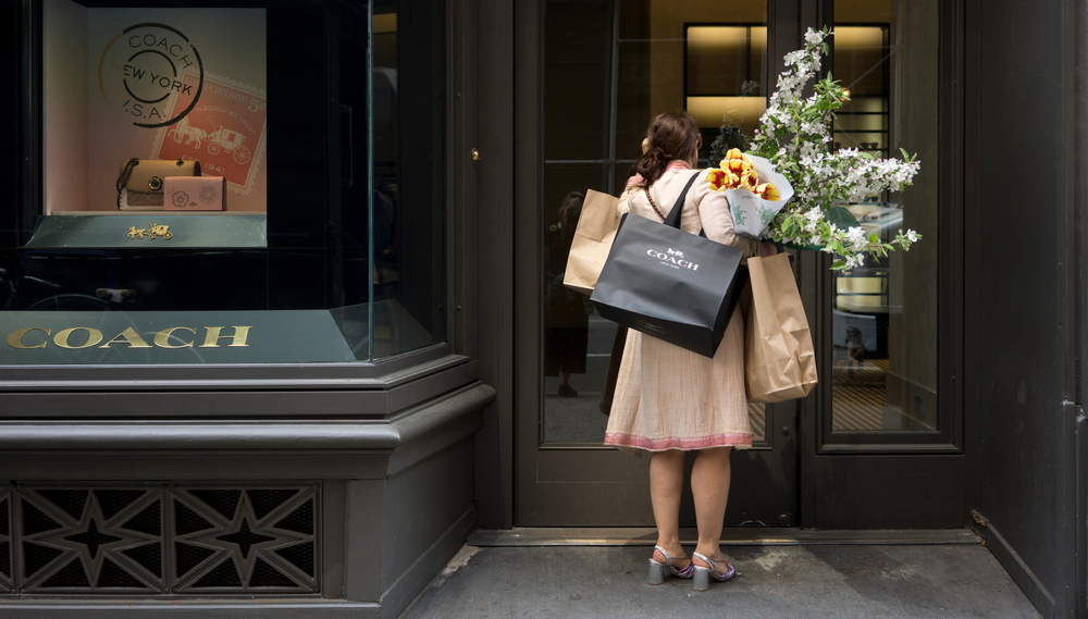 A woman carrying three shopping bags and two bouquets of flowers stands outside the Coach store facing the door, with her back to the camera