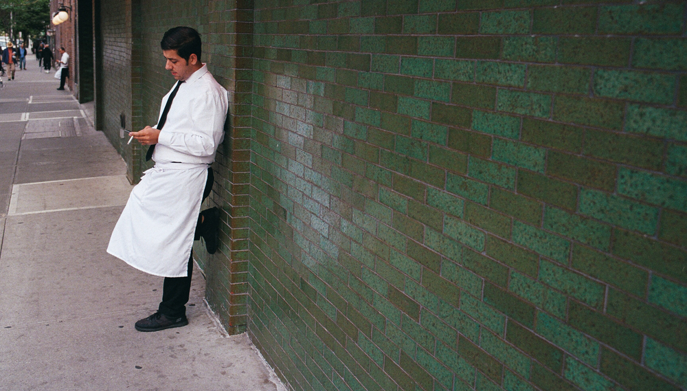 A waiter stands outside resting against a wall and taking his smoke break