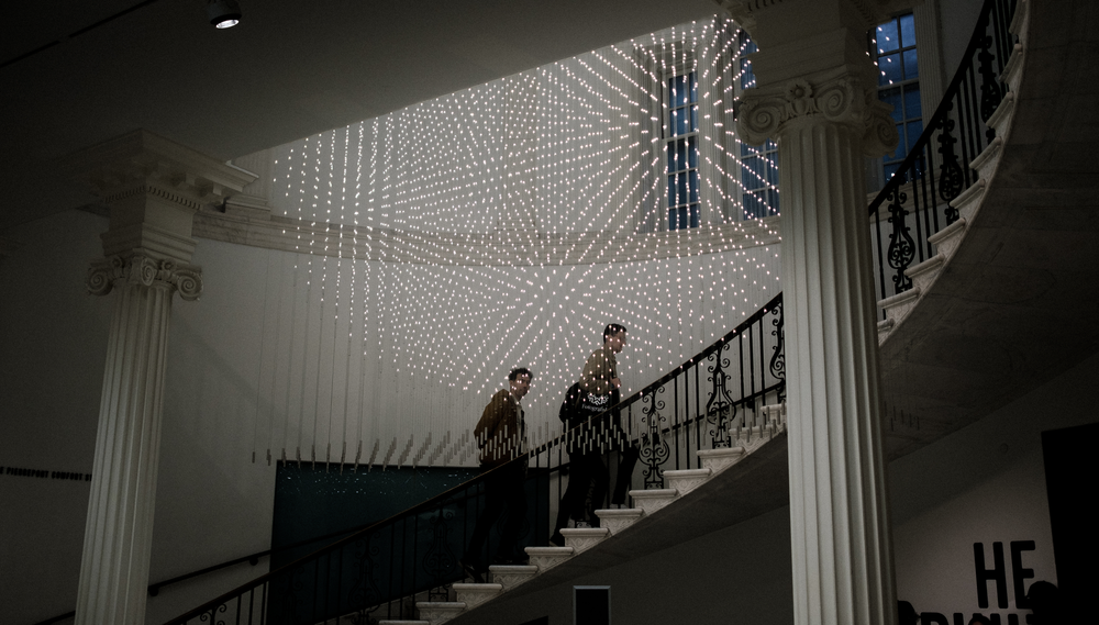 Two people walk up a spiral staircase, above which a light installation is projected on the ceiling