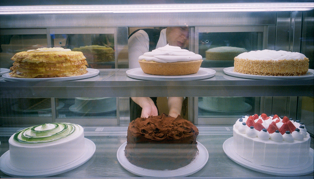 A bakery employee removes a chocolate cake from a display case displaying six ornate cakes on two shelves