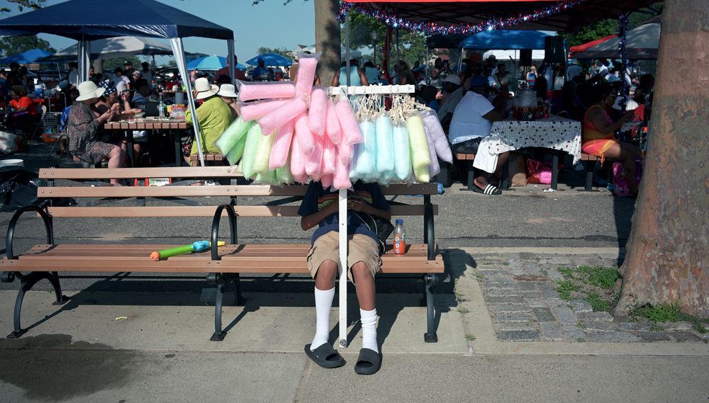 A boy sits on a bench selling cotton candy of various colors from a rack while people dine at picnic tables under tarps behind hi