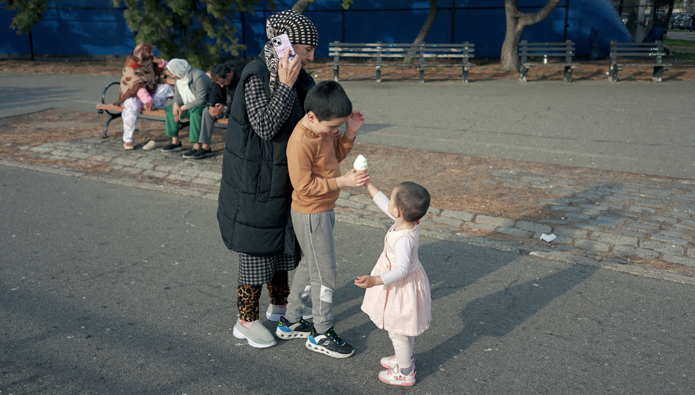 An elementary school boy hands an ice cream cone to his toddler sister while their mother stands next to them taking a phone call
