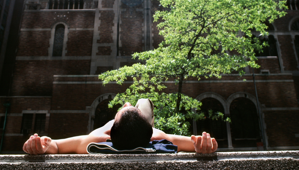 A man sunbathes on the ground outside a building in front of a tree