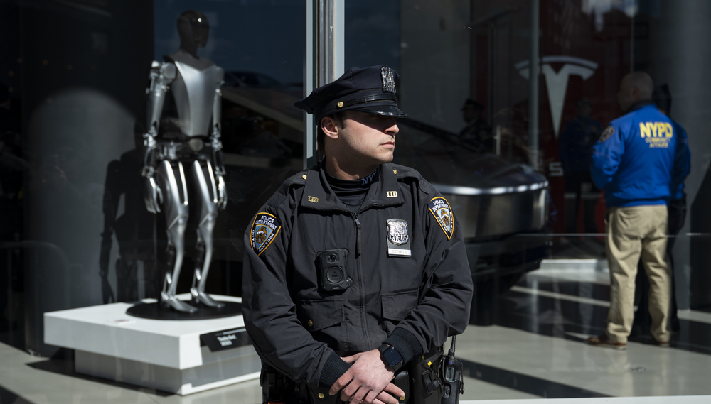 An officer from the New York Police department stands guard outside of a Tesla store window displaying a robot while another officer stands in the back of the frame investigating inside with 
