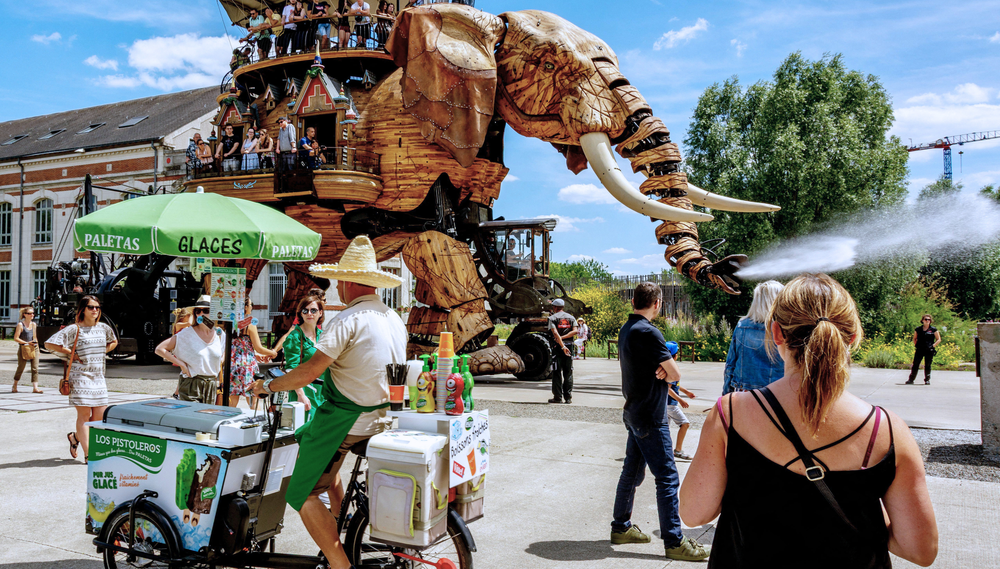A paletas vendor and several tourists move through a street in Nantes as the Great Elephant mechanical art installation walks past them