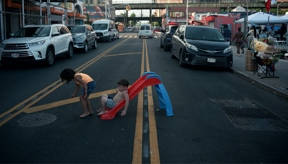 Two children play on a toy slide in the middle of a road lined with cars parked on the curb