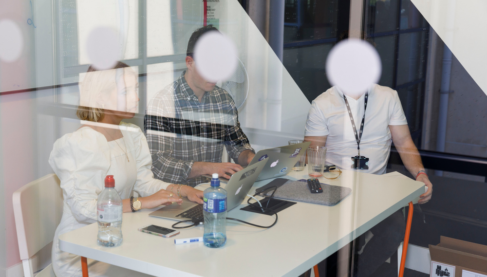 Three people working on their laptops gather at a table in their office behind a glass wall with a row of four white dots imprinted on it; the dots obscure their faces