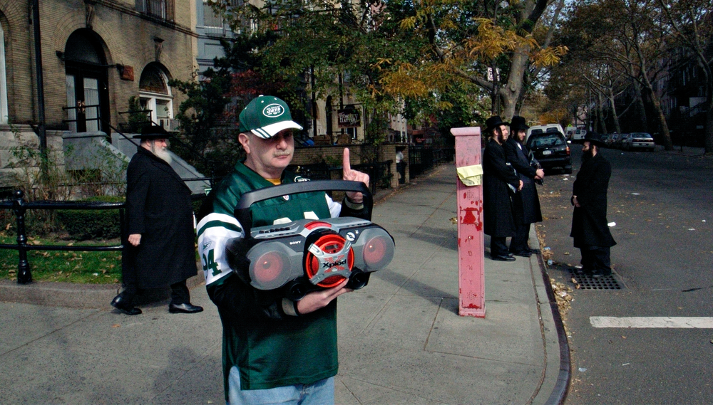 A man in a sports jersey stands at a street corner and holds up a radio while a group of Hasidic men gather on the sidewalk behind him