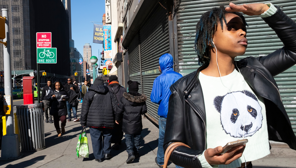 A woman wearing a shirt depicting a panda stands at the corner of a busy sidewalk, looking into the distance and holding her hand above her brow to block the sun