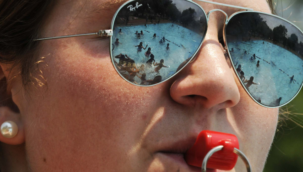 A close-up of a lifeguard with a whistle in her mouth whose sunglasses reflect the image of a pool full of swimmers
