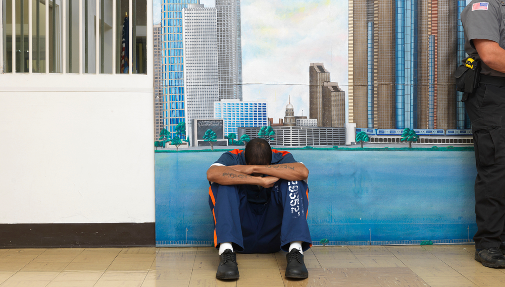 An inmate at the Michigan Reformatory sits crouched with his head bent down in his arms in front of a painted backdrop of a city and a window with bars; a corrections officer is seen standin