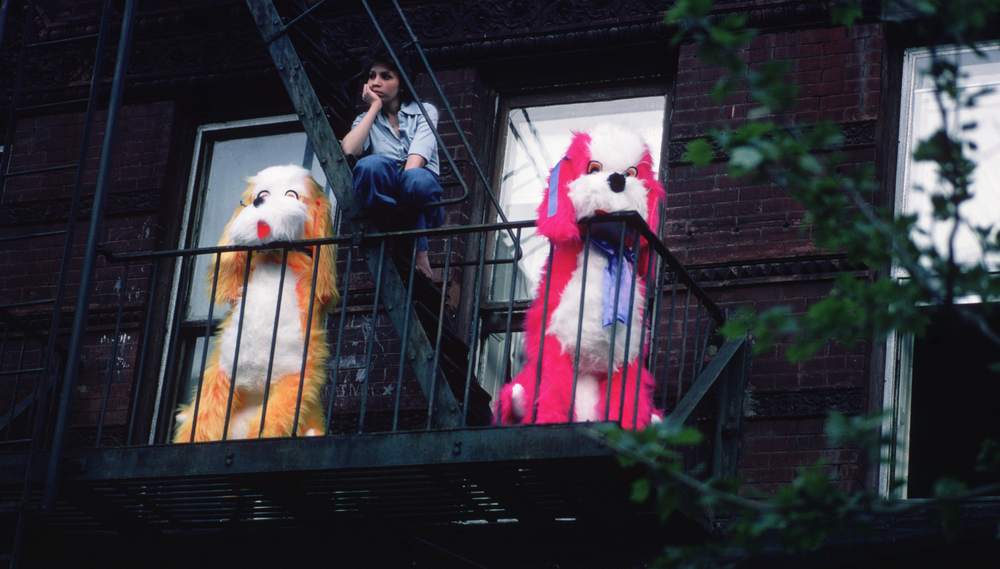 A woman sits on her fire escape next to two giant stuffed dogs, one orange and one hot pink
