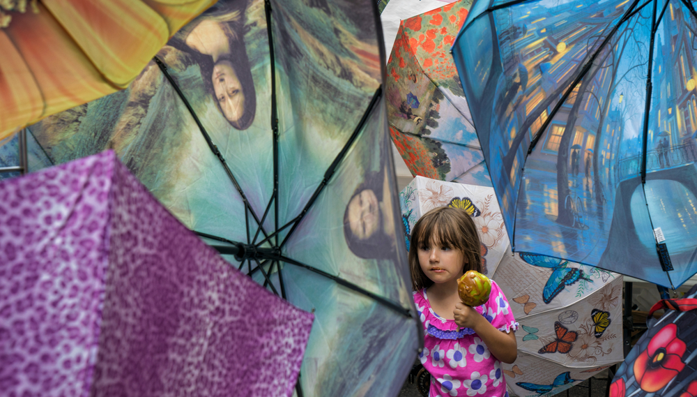 A young girl holding a caramel apple stands amidst a display of umbrellas featuring famous artworks and colorful patterns