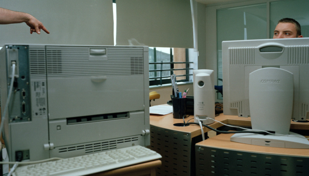 An office worker sits behind his computer monitor in the right of frame; in the left of frame, an unseen figure points a finger towards his desk above another monitor