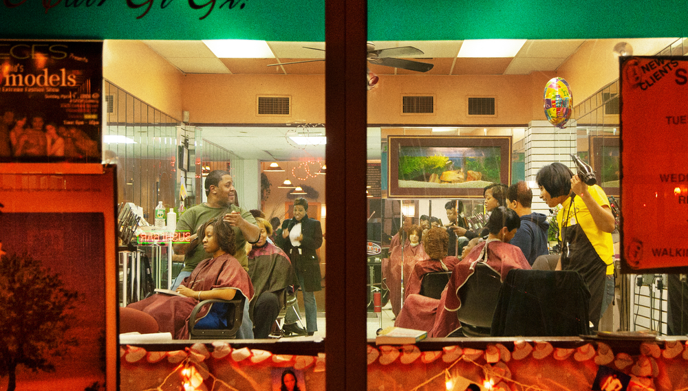 The patrons and employees of a hair salon are seen through the window, the edges of which are papered with flyers and decoration