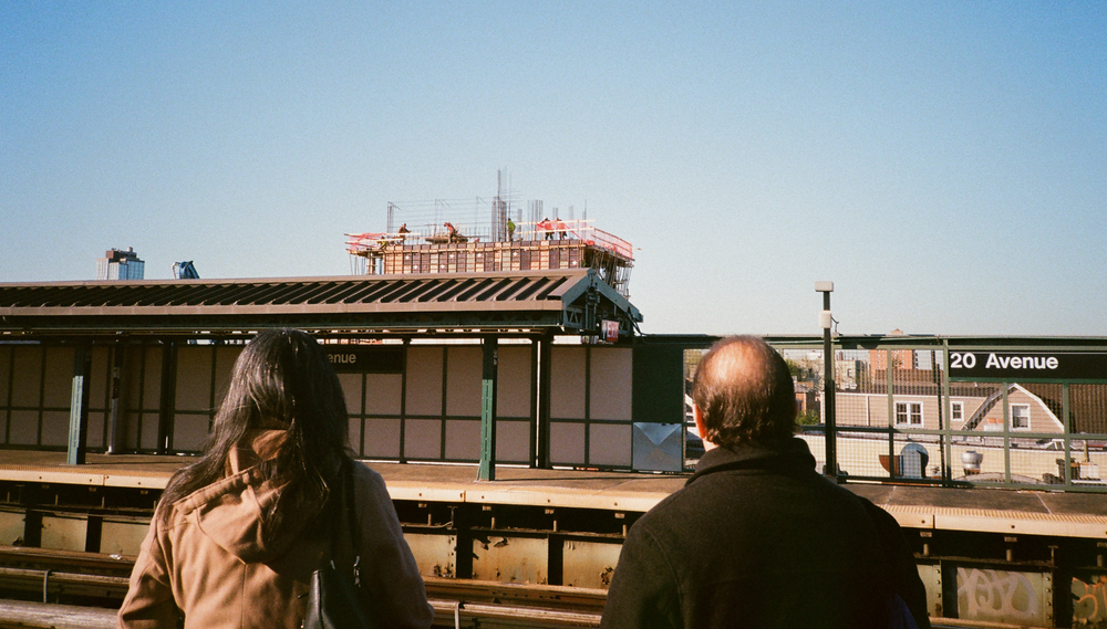 Two people stand on the platform of the above-ground 20 Avenue subway station looking at a high-rise apartment being constructed in the distance
