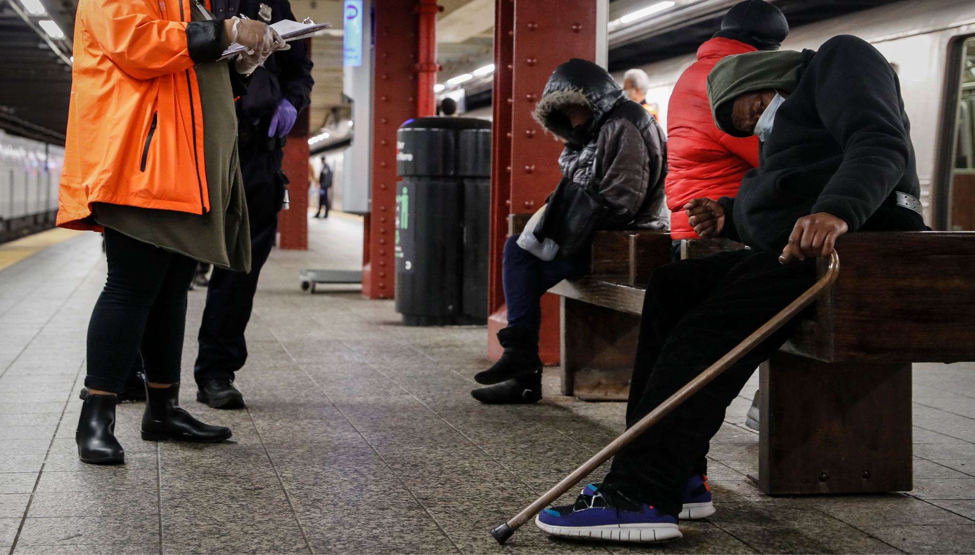 A police officer and a homeless outreach worker assist a homeless man sleeping on a bench on a Subway platform