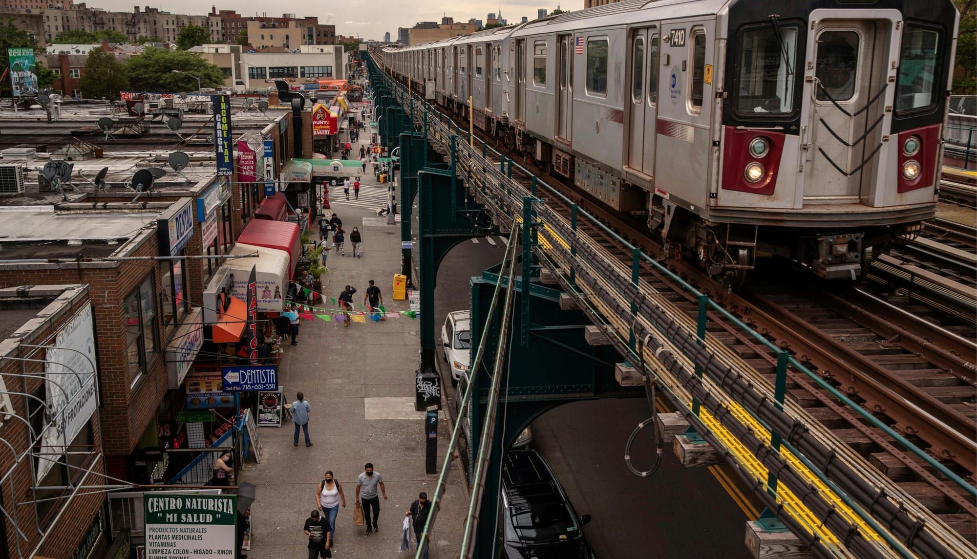 A train on an elevated subway line passes shops and pedestrians on Roosevelt Avenue in Corona, Queens