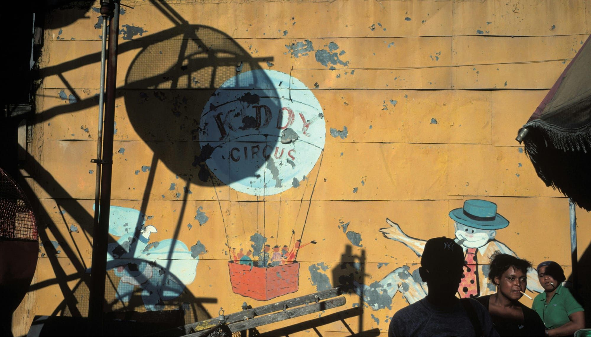 Shadows of people and infrastructure are cast upon a mural depicting a circus