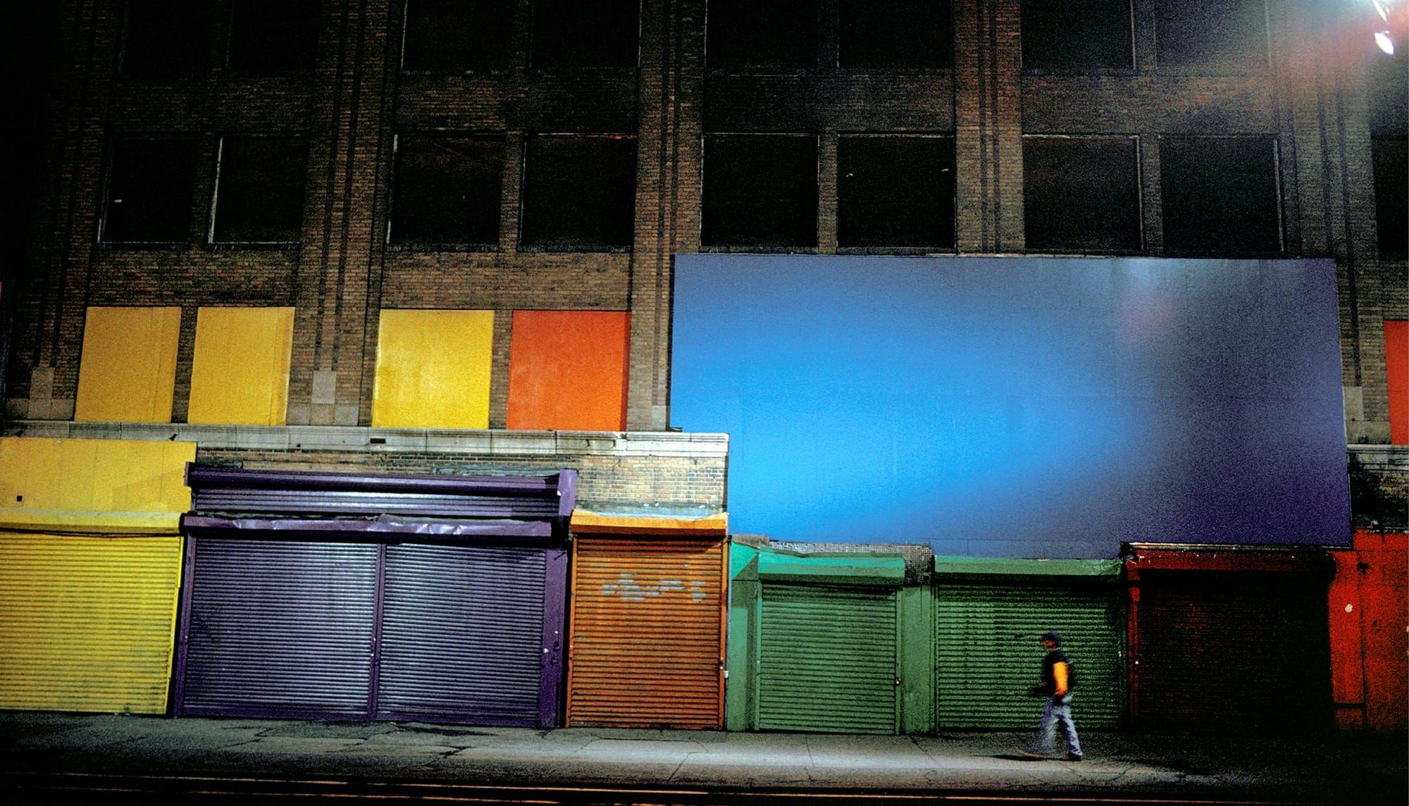A man walks in front of a row of buildings with some windows boarded up with colorful boards and their doors painted a variety of bright colors