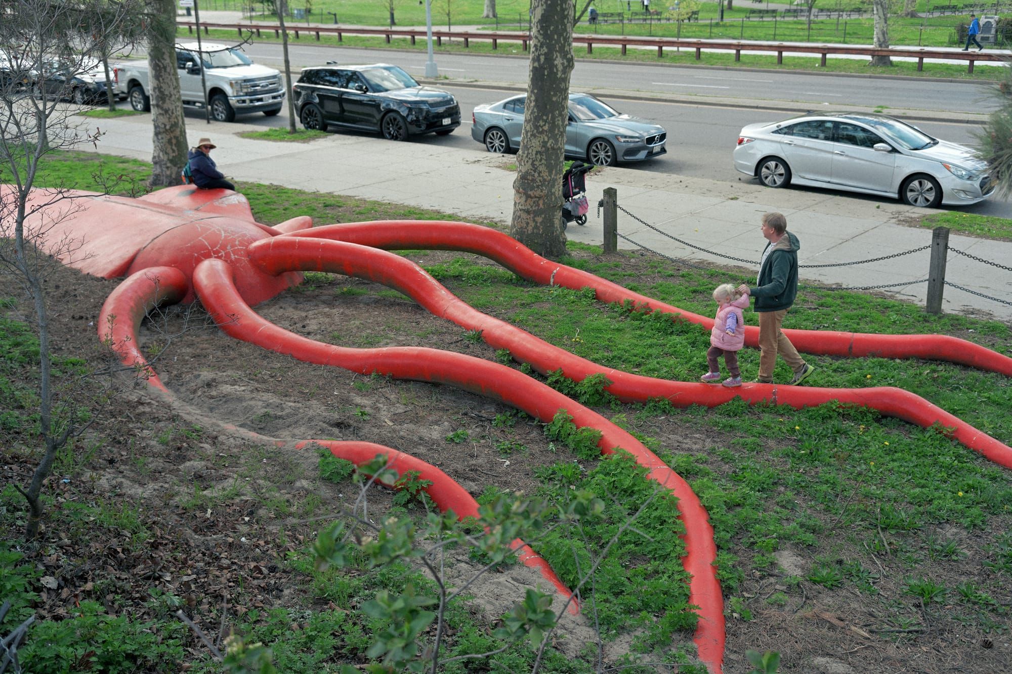 A toddler's father walks with her next to a large sculpture of a squid at a park to meet her mother on the other side