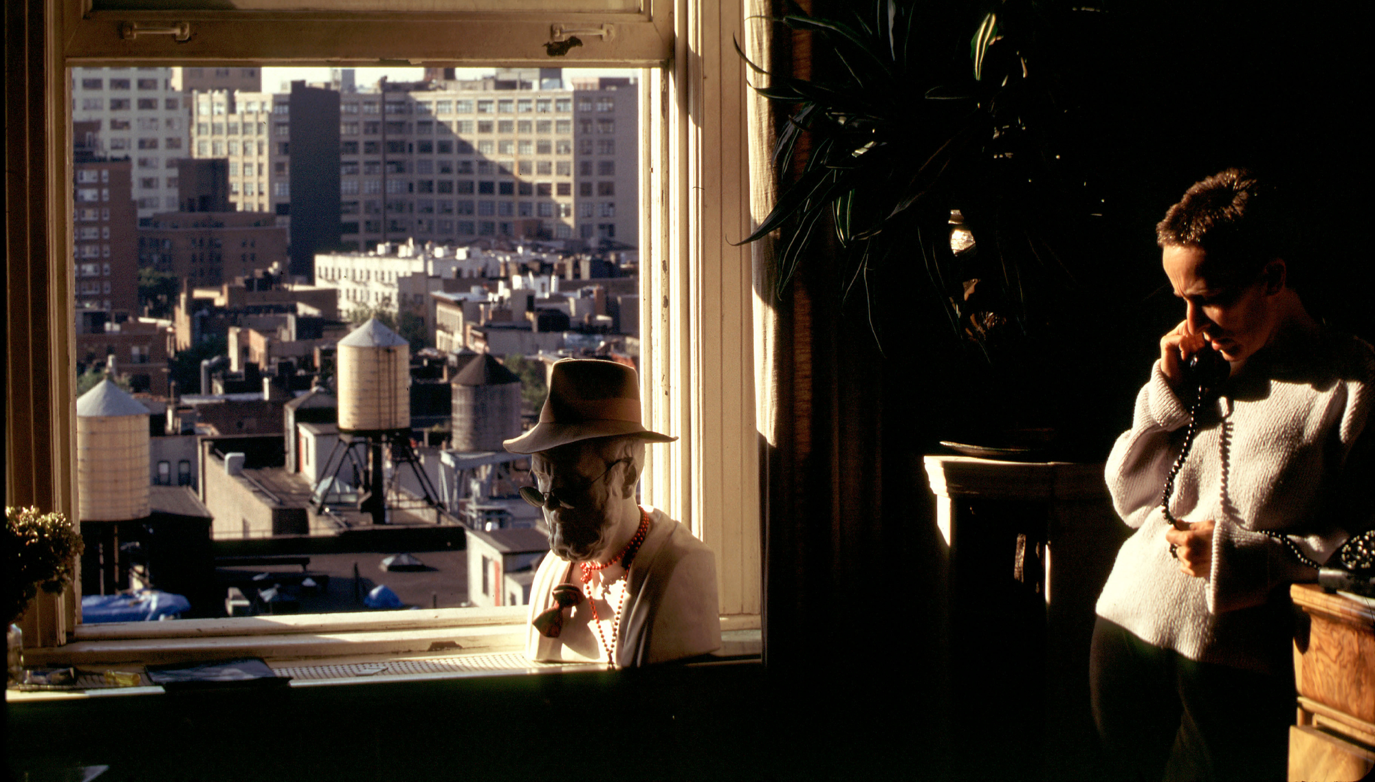 A person stands next to a window in their apartment taking a phone call; outside the window can be seen water tanks on rooftops