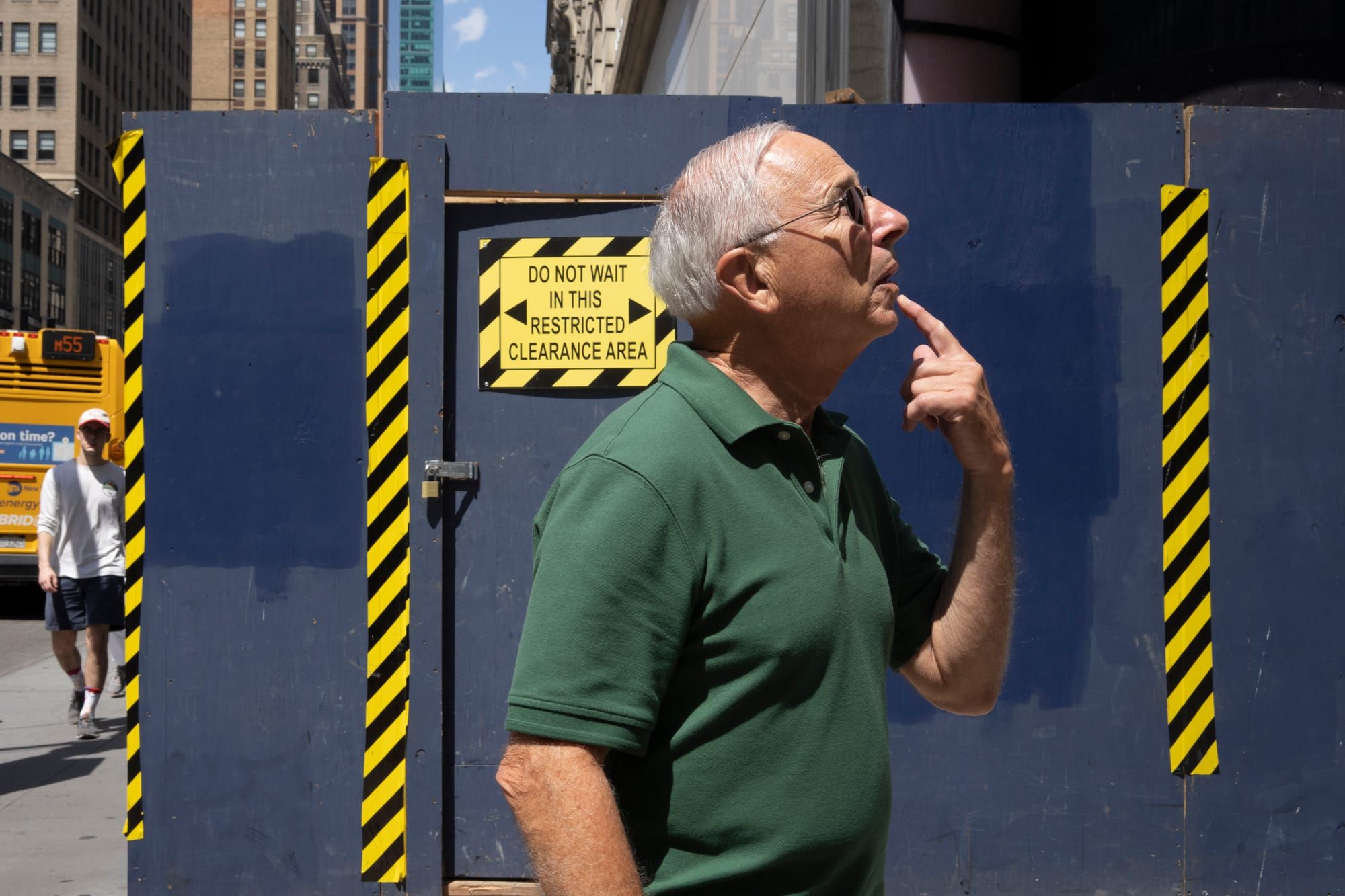 A man standing in front of a construction site holds his index finger to his chin and looks upward