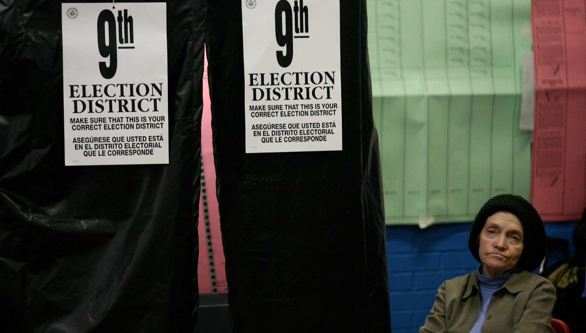 A woman wearing a knit hat sits next to a voting booth displaying signs indicating it is for voters in the 9th election district