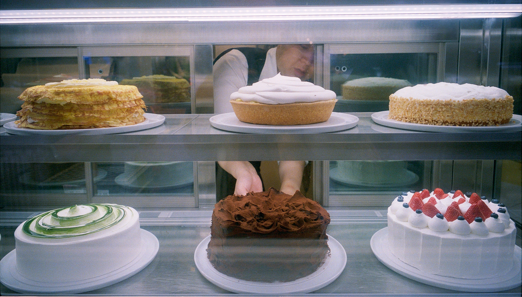 A bakery employee removes a chocolate cake from a display case displaying six ornate cakes on two shelves