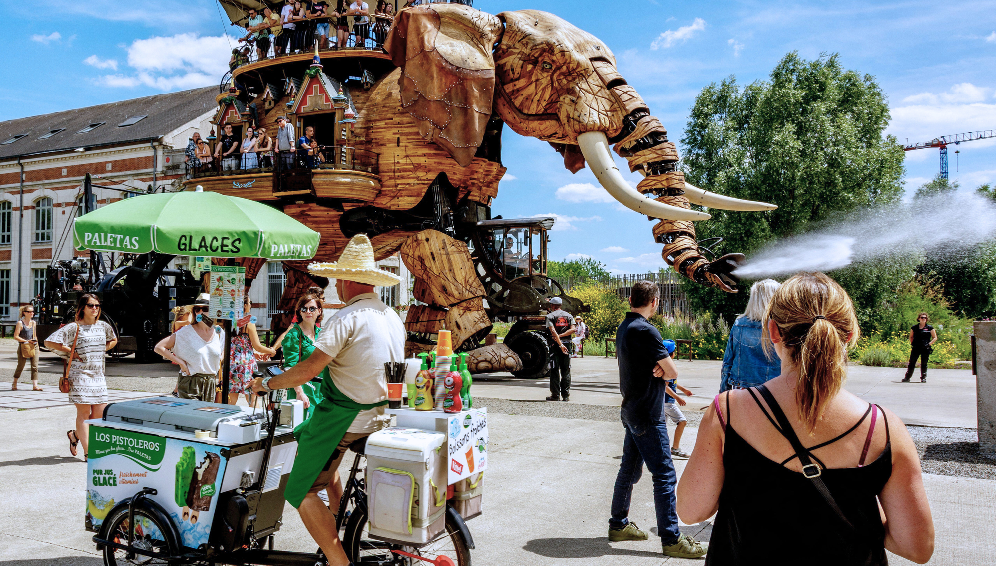 A paletas vendor and several tourists move through a street in Nantes as the Great Elephant mechanical art installation walks past them