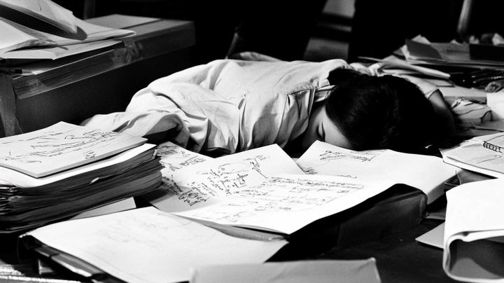 Image in black and white of a female asleep on a desk with books and notes all around