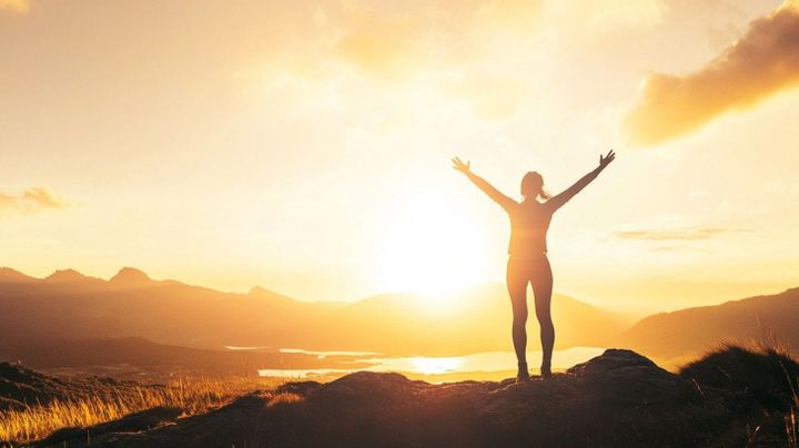 Image of a woman on a mountaintop in a celebratory pose with a sunset backdrop
