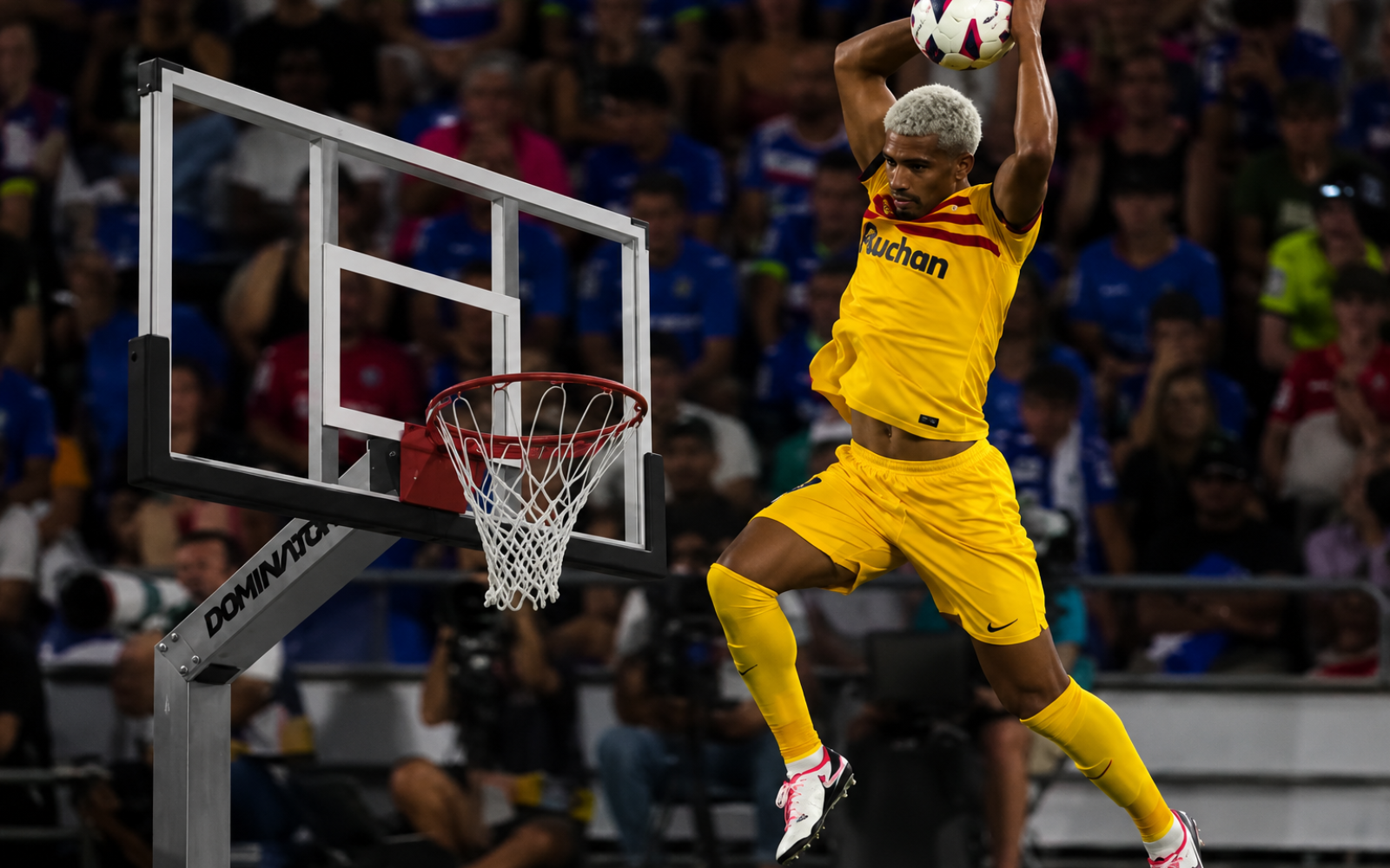 Footballer Araujo in a yellow kit leaps beside a basketball hoop with a football raised above his head in a packed stadium.