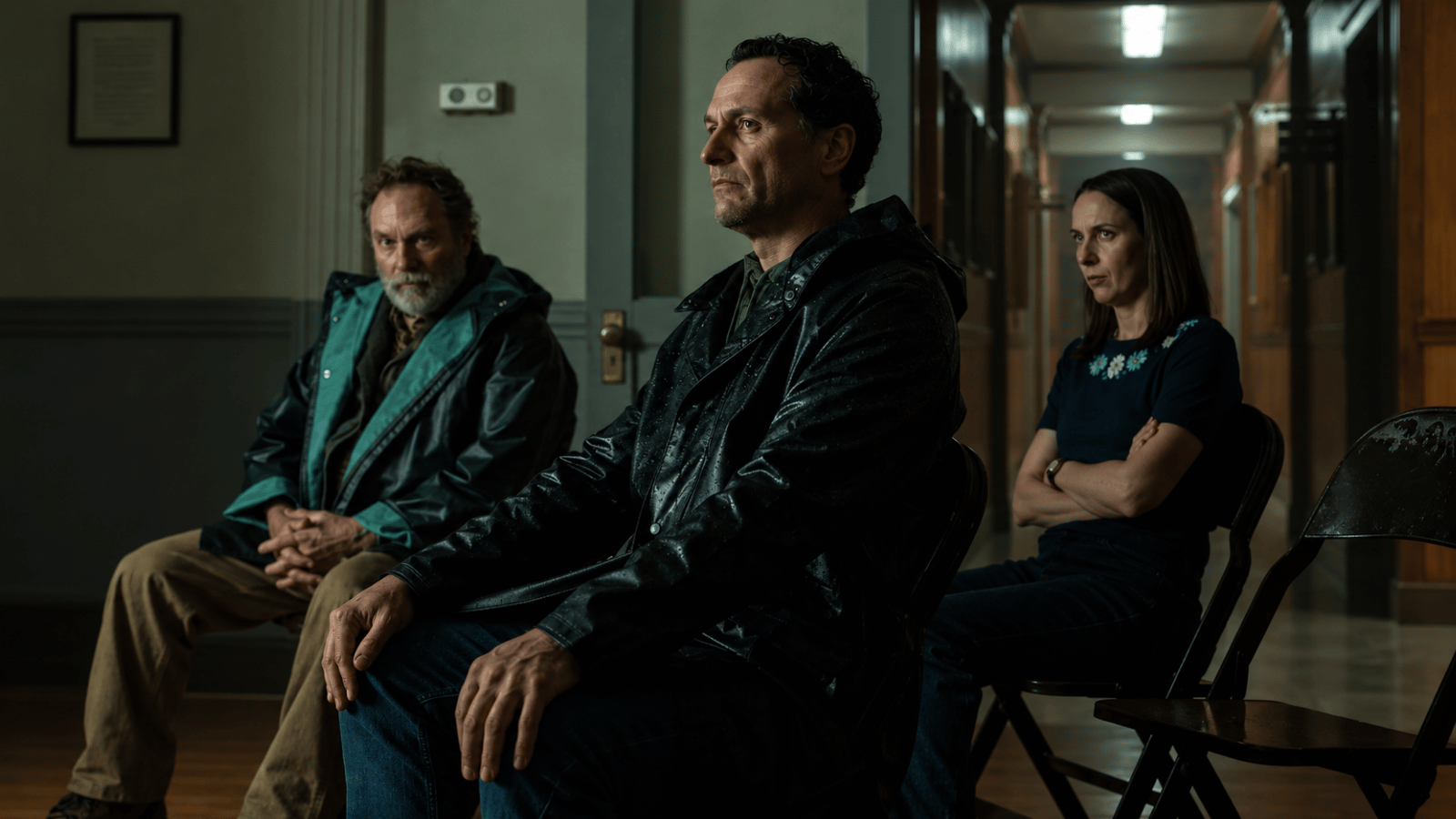 Three tense characters sit on folding chairs in a dim institutional hallway.