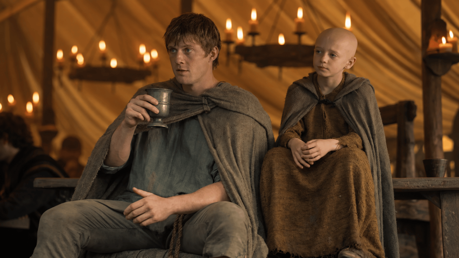 A young knight holding a metal cup sits beside a bald boy inside a candlelit medieval hall.