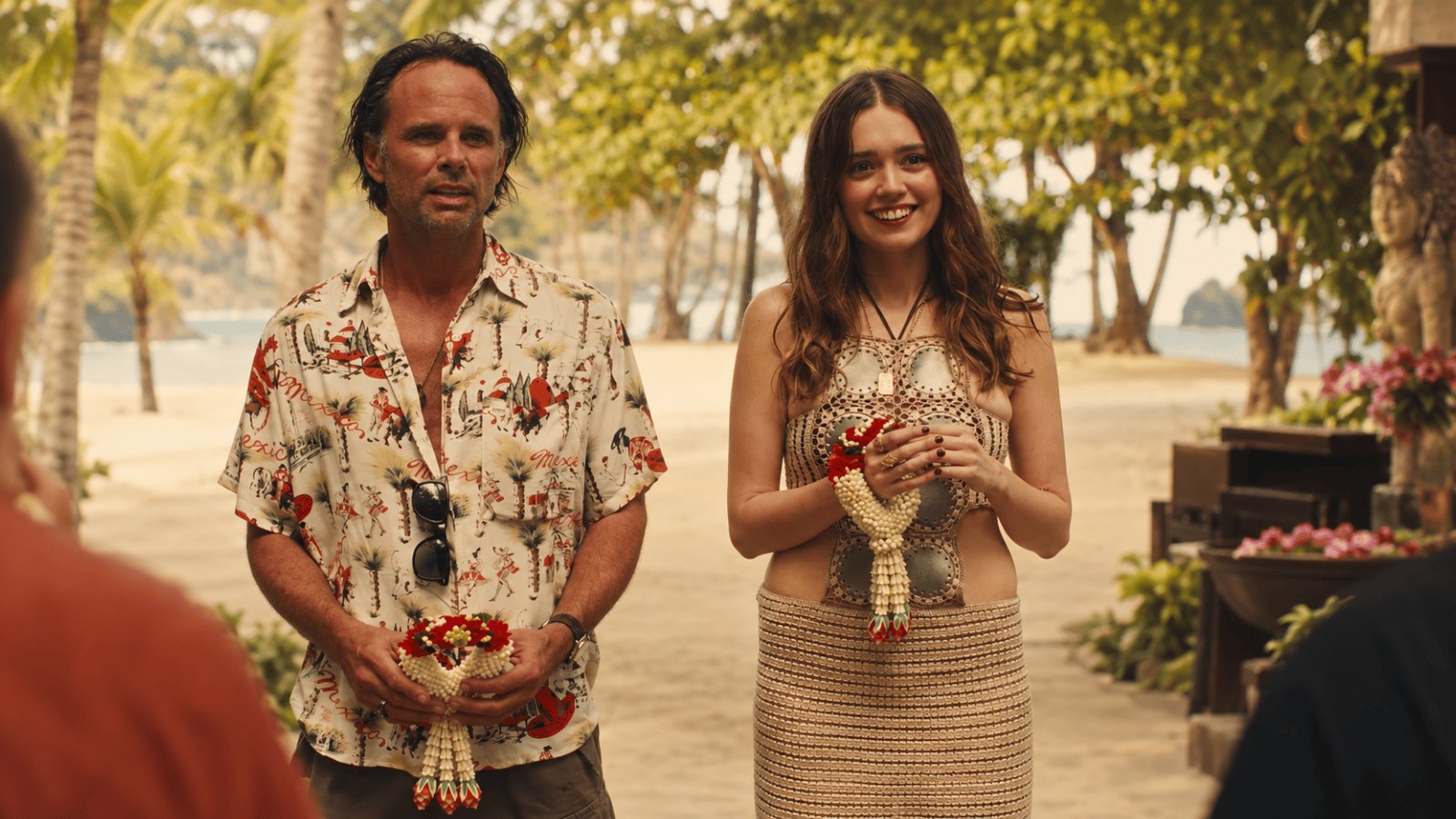 A man and young woman holding flower garlands welcome guests at a tropical beach resort.