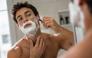 Man shaving his face with a razor in front of a bathroom mirror using proper technique.