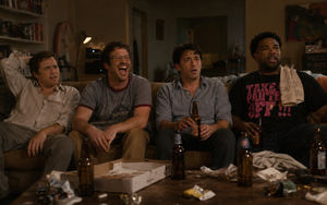 Four friends sit on a worn sofa watching a film in a dimly lit living room, with beer bottles, an empty pizza box and snacks scattered across a cluttered coffee table.