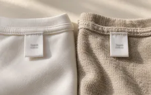Macro photograph of two identical white 100% Cotton fabric tags, each sewn onto a different t-shirt fabric - one smooth, tightly woven and dense, the other loosely knit and fuzzy