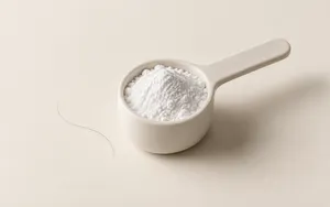 A single scoop of creatine powder resting beside one undisturbed strand of hair on a soft ivory surface.