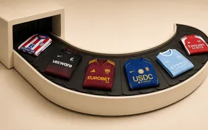 2026/27 football shirts from Chelsea, Manchester City, Arsenal, Roma, Atlético Madrid and Freiburg moving around a cream airport-style baggage carousel.