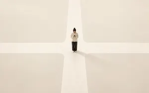 A person stands at the centre of an empty cream-coloured crossroads, looking down at a phone held in both hands.