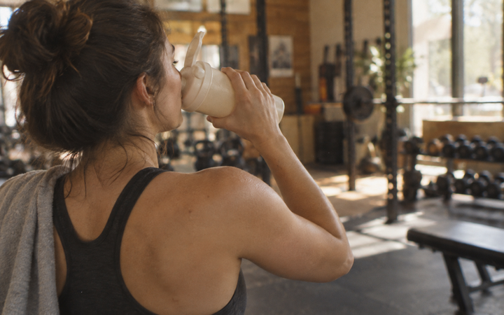 Woman drinking from a plain cream shaker bottle in a softly lit gym, photographed from behind with weights and benches blurred in the background.