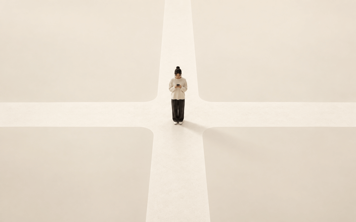 A person stands at the centre of an empty cream-coloured crossroads, looking down at a phone held in both hands.